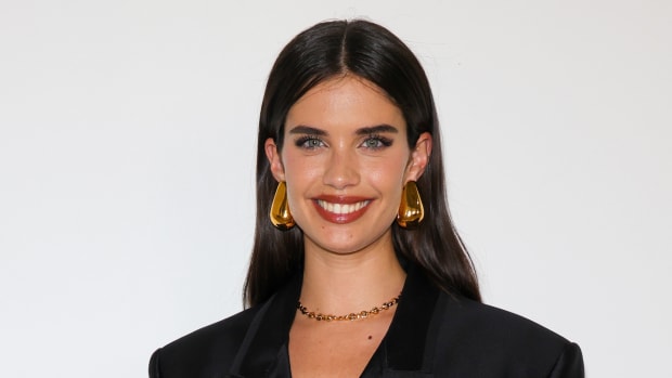 Sara Sampaio poses in front of a white backdrop in a black blazer, red lip and oversized gold earrings.